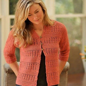 May include: A coral-colored, open-front crochet cardigan featuring three-quarter length sleeves and a V-neckline. The garment is constructed with alternating bands of solid crochet stitches and openwork patterns, secured by three small, round, neutral-toned buttons.