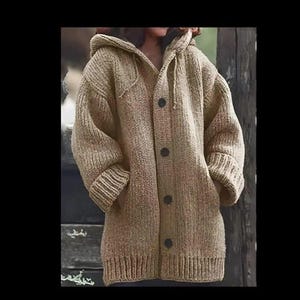 Op de afbeelding: Een beige vest met capuchon, grove breistructuur, lange mouwen met geribbelde boorden en een knoopsluiting aan de voorzijde met vier donkere, ronde knopen. Het kledingstuk is voorzien van een geribbelde zoom en twee zijzakken voor extra detail.