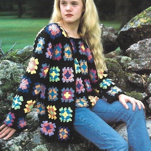 Op de afbeelding: Een zwart gehaakt vest en bijpassende top met een kleurrijk granny square patroon in tinten blauw, geel, roze en paars. Het vest met lange mouwen heeft een ronde halslijn en wordt gecombineerd met een blauwe spijkerbroek met vetersluiting aan de zijkant.