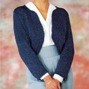Könnte beinhalten: Eine dunkelblaue Strickjacke mit langen Ärmeln und strukturierter Oberfläche. Das Kleidungsstück hat einen abgerundeten Ausschnitt und einen dekorativen Picot-Rand entlang der Vorderöffnung und an den Ärmelbündchen. Sie wird über einem weißen Hemd getragen.