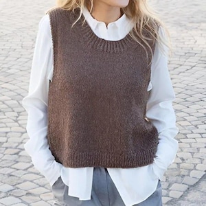 May include: A dark brown sleeveless knit sweater vest featuring a ribbed crew neckline and a ribbed hem. The vest is layered over a crisp white long-sleeved button-down shirt, which is tucked into a pair of light gray trousers.