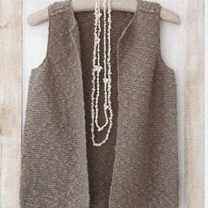 May include: A sleeveless, open-front waistcoat in a textured, medium-brown knit fabric. The waistcoat is displayed on a light-coloured wooden hanger. A long, multi-strand necklace made of small, white, natural seashells hangs down the centre of the waistcoat.