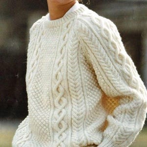Könnte beinhalten: Ein cremeweißer Langarmpullover mit einem komplexen Strickmuster aus vertikalen Zopfmustern, geflochtenen Texturen und kleinen erhabenen Noppendetails. Das Kleidungsstück hat einen gerippten Rundhalsausschnitt und gerippte Bündchen aus dickem Garn.