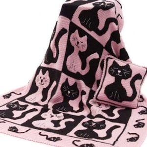 Op de afbeelding: Een gebreide deken en een bijpassend vierkant kussen met een herhalend patroon van zwarte katten op een lichtroze achtergrond en lichtroze katten op een zwarte achtergrond. De rand van de deken toont een rij kleine zwarte katsilhouetten.