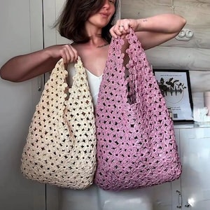 May include: Two crochet tote bags made from joined flower motifs. One bag is a light cream color, and the other is a dusty rose pink. Both bags feature an openwork design with integrated handles, showcasing a repeating floral pattern throughout.