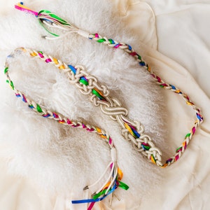 May include: A braided belt with colorful ribbons. The belt is made of white and gold rope, with sections of braided rainbow-colored ribbons. The belt has a decorative knot in the center and tassels at the end.