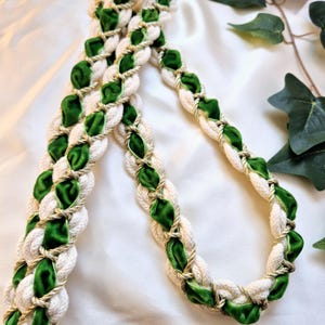 May include: A braided necklace featuring cream-colored rope intertwined with vibrant green velvet and gold-colored metallic thread. The necklace is displayed on a white satin surface, with green ivy leaves in the background.