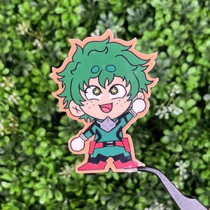 May include: A colorful sticker featuring a cartoon character with green hair and a green and red outfit. The character has a cheerful expression with freckles and is waving. The sticker is held by tweezers against a backdrop of green foliage.