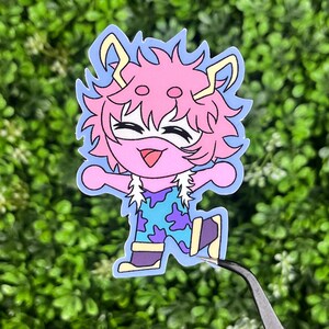 May include: A sticker featuring a cartoon character with pink hair and a light blue outline. The character is smiling and wearing a blue and purple outfit. The sticker is held by tweezers against a green background.