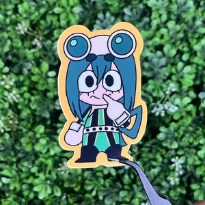 May include: A sticker featuring a cartoon character with teal hair, large eyes, and a frog-like appearance. The character is wearing a white and green outfit with black accents. The sticker has a yellow border and is held by tweezers.