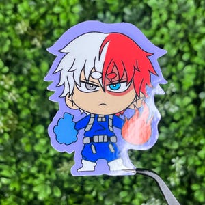 May include: A sticker featuring a cartoon character with white and red hair, blue eyes, and a stern expression. The character wears a blue outfit and has a blue glove on one hand and a flame on the other. The sticker has a purple outline.