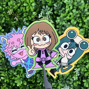 May include: Three colorful cartoon character stickers. One is pink with spiky hair, another is brown-haired with a peace sign, and the third is green with goggles. The stickers are on a green leafy background.