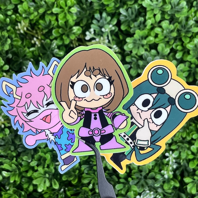 May include: Three colorful cartoon character stickers. One is pink with spiky hair, another is brown-haired with a peace sign, and the third is green with goggles. The stickers are on a green leafy background.