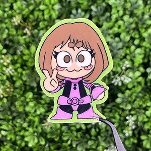 May include: A colorful sticker featuring a cartoon character with brown hair and large eyes. The character is wearing a pink and black outfit and making a peace sign with their hand. The sticker has a green border and is set against a green background.