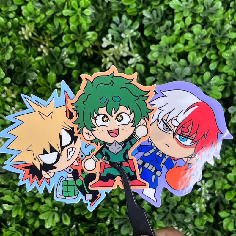 May include: Three colorful stickers featuring anime characters. The stickers depict characters with spiky hair, green hair, and red and white hair. The characters are wearing superhero-style outfits. The stickers are on a green, leafy background.