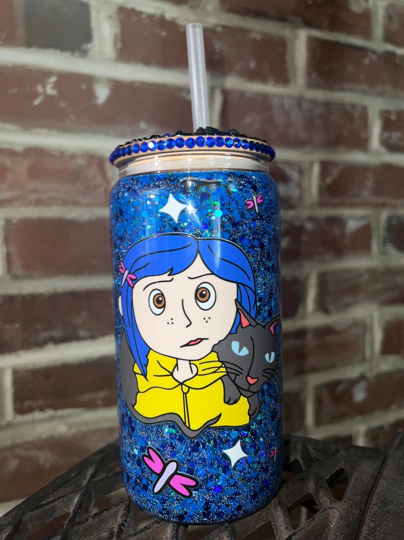 May include: A blue and black glitter tumbler with a blue rhinestone rim. The tumbler features a cartoon image of a girl with blue hair and a black cat. The girl is wearing a yellow jacket. The tumbler has a clear straw.