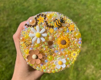 May include: A round, clear resin coaster with a bee, flowers, and pearls. The coaster is decorated with yellow, white, brown, and gold accents.
