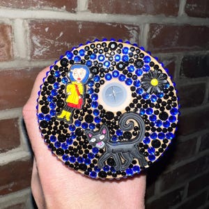 May include: A round wooden coaster with a black cat, a girl in a yellow raincoat, and a black flower with a yellow center. The coaster is covered in black and blue rhinestones.
