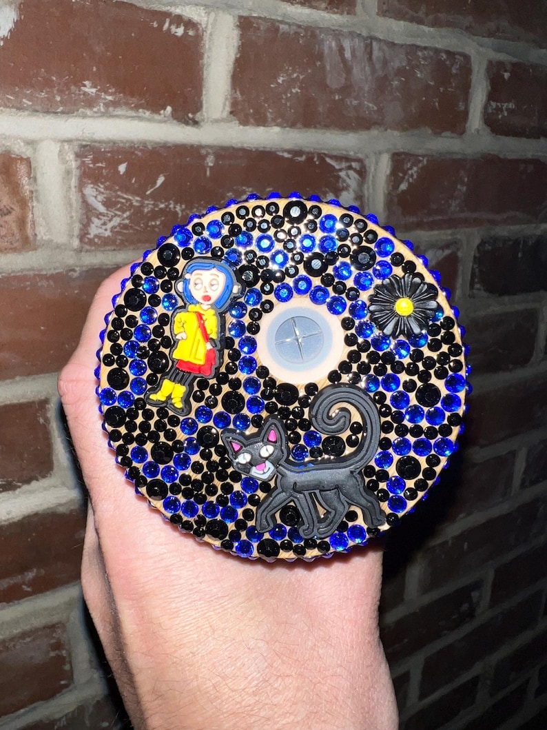 May include: A round wooden coaster with a black cat, a girl in a yellow raincoat, and a black flower with a yellow center. The coaster is covered in black and blue rhinestones.