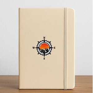 May include: A beige notebook with an elastic band closure. The cover features a black and white compass design with a mountain and sunset scene. The compass points to North, South, East, and West.