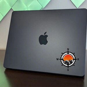 May include: A black laptop with a compass sticker featuring a mountain range and sunset design. The sticker is centered on the laptop's lid. The laptop has a silver apple logo.