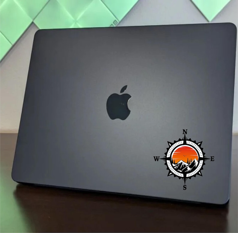 May include: A black laptop with a compass sticker featuring a mountain range and sunset design. The sticker is centered on the laptop's lid. The laptop has a silver apple logo.