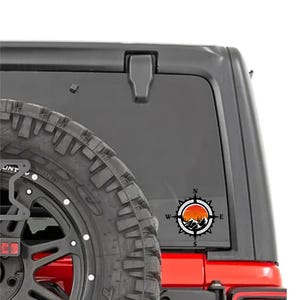 May include: A black and orange compass sticker with a mountain and sunset design. The sticker is affixed to the rear window of a black vehicle, near a spare tire. The compass points North, South, East, and West.