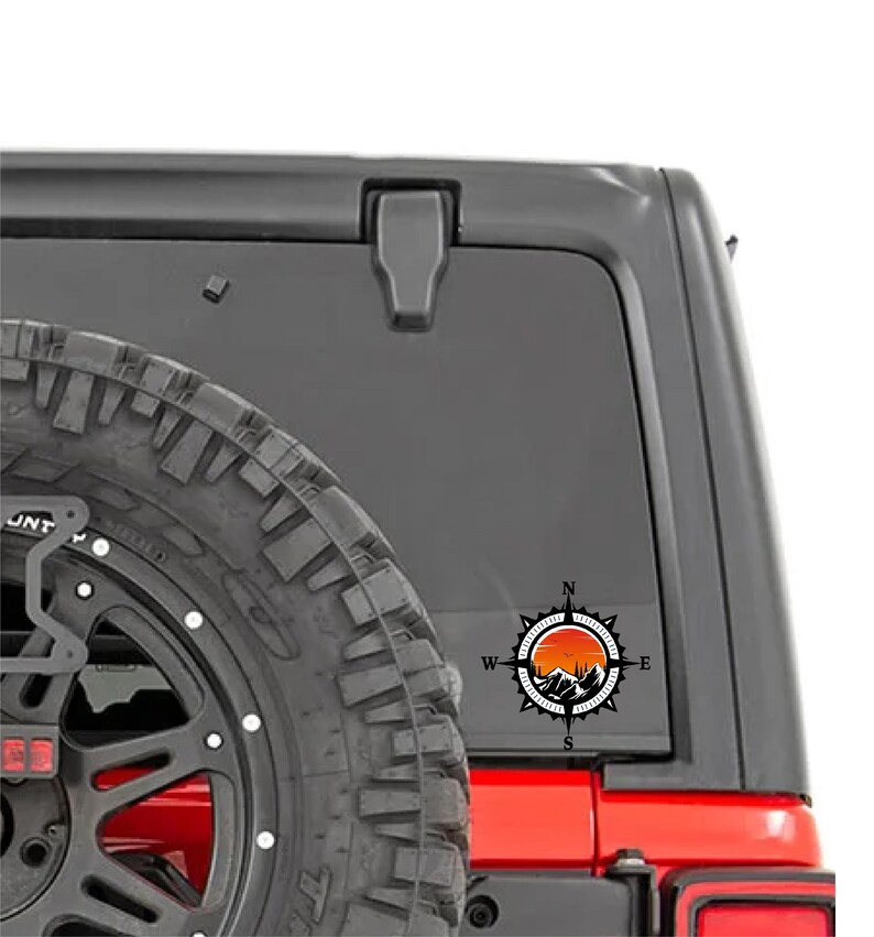 May include: A black and orange compass sticker with a mountain and sunset design. The sticker is affixed to the rear window of a black vehicle, near a spare tire. The compass points North, South, East, and West.