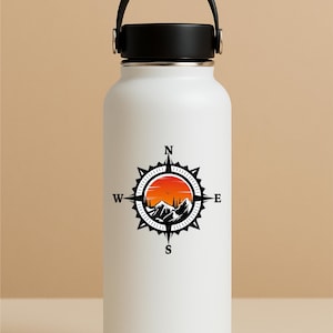 May include: White insulated water bottle with a black handle and lid. The bottle features a black and orange compass design with mountains, trees, and a sunset. The compass points to North, South, East, and West.