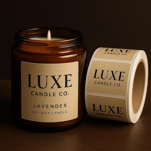 May include: A lit soy wax candle in an amber glass jar with a beige label reading LUXE CANDLE CO. LAVENDER SOY WAX CANDLE, positioned next to a roll of matching beige adhesive labels featuring the text LUXE CANDLE CO. and HAND CRAFTED.