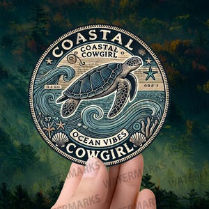 May include: A round sticker with a sea turtle design. The sticker has the words "Coastal Cowgirl" and "Ocean Vibes" with images of waves, starfish, and seashells. The sticker is held in a hand.