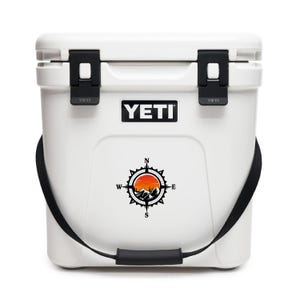 May include: White YETI cooler with black latches, handle, and strap. The cooler features a compass design with a mountain scene and the YETI logo. The cooler is designed to keep items cold.