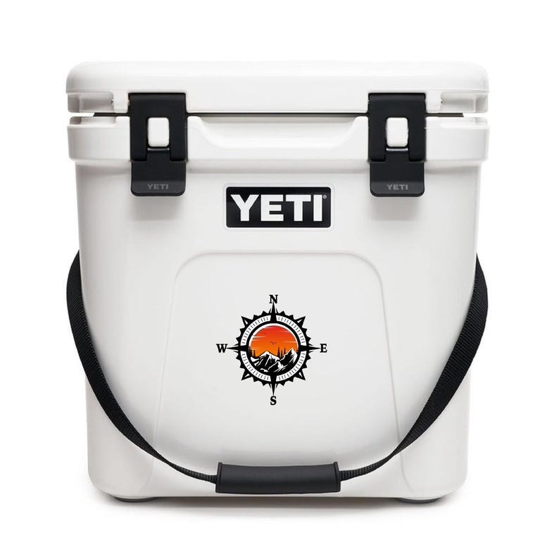 May include: White YETI cooler with black latches, handle, and strap. The cooler features a compass design with a mountain scene and the YETI logo. The cooler is designed to keep items cold.