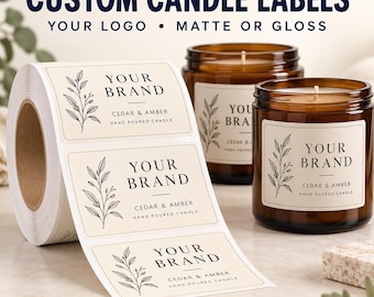 May include: A roll of rectangular adhesive labels with a cream background, black text reading YOUR BRAND and CEDAR & AMBER HAND POURED CANDLE, and a black line drawing of a leafy branch. Two amber glass candle jars feature the same label design.