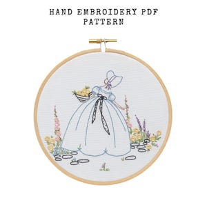 May include: Hand embroidery PDF pattern featuring a woman in a blue dress and bonnet, carrying a basket of yellow flowers. The design is set within a wooden embroidery hoop, with floral accents and the text "HAND EMBROIDERY PDF PATTERN" at the top.