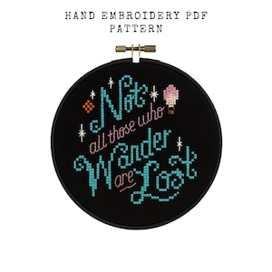 May include: A black embroidery hoop with the text "Not all those who wander are lost" in teal and pink cross-stitch. The words are surrounded by stars and a hot air balloon. The text "HAND EMBROIDERY PDF PATTERN" is above the hoop.