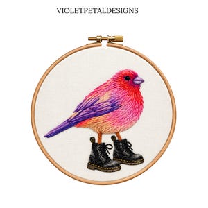 May include: An embroidery hoop containing a white fabric panel with the text VIOLETPETALDESIGNS at the top. The embroidery depicts a vibrant pink, red, and purple bird wearing a pair of black lace-up combat boots on its feet.