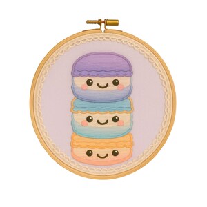 Peut inclure: Broderie sur un cercle en bois représentant une pile de trois macarons colorés. Les macarons souriants sont violets, bleus et oranges, sur un fond lilas. Le cercle en bois est orné d'une bordure en dentelle, mesurant environ 15 cm de diamètre.