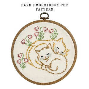 May include: Hand embroidery PDF pattern featuring two sleeping cats nestled together, surrounded by pink flowers. The design is stitched on a cream-colored fabric and framed in a wooden hoop. The text "HAND EMBROIDERY PDF PATTERN" is at the top.