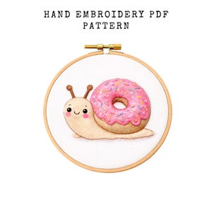 May include: Hand embroidery PDF pattern featuring a cute snail with a donut shell. The snail is beige with a pink donut shell covered in sprinkles. The embroidery is in a wooden hoop, with the text "HAND EMBROIDERY PDF PATTERN" above.
