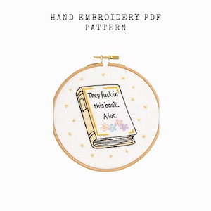 May include: Hand embroidery PDF pattern featuring a book design with the text "They fuck in this book. A lot." The book is cream-colored with gold trim and floral accents, set in a wooden embroidery hoop.