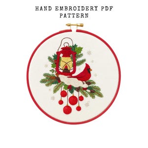 May include: Hand embroidery PDF pattern featuring a red cardinal, lantern, and ornaments. The design is set in a round embroidery hoop with a red border. The text "HAND EMBROIDERY PDF PATTERN" is at the top.