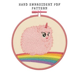 May include: Hand embroidery PDF pattern featuring a pink, fluffy unicorn with a rainbow. The unicorn has a horn and is embroidered on a cream-colored fabric, set in a pink embroidery hoop. The text "HAND EMBROIDERY PDF PATTERN" is at the top.