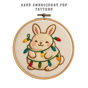 以下が含まれることがあります： 手刺繍PDFパターン。カラフルなクリスマスライトで飾られた、笑顔のベージュ色のウサギが描かれています。デザインは木製の刺繍枠に収められており、上部には「HAND EMBROIDERY PDF PATTERN」の文字があります。生地はクリーム色です。