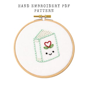 May include: A hand embroidery pattern featuring a smiling book with a red heart and green leaves. The design is stitched on white fabric and framed in a wooden embroidery hoop. The text "HAND EMBROIDERY PDF PATTERN" is at the top.