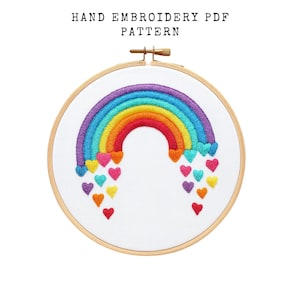 May include: Hand embroidery PDF pattern featuring a colorful rainbow design with heart accents. The rainbow arcs in shades of purple, blue, green, yellow, and red. The design is set in a wooden embroidery hoop.