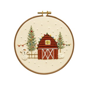 May include: Embroidered hoop art featuring a red barn with a white cross design on the doors, a yellow window, and string lights. Two green Christmas trees flank the barn, and a snowman stands near a small fence. Snowflakes are scattered across the cream-colored background.