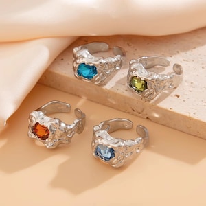 Puede incluir: Cuatro anillos de plata con piedras preciosas de diferentes colores. Los anillos tienen un diseño texturizado y orgánico. Las piedras preciosas son azules, verdes, naranjas y azul claro.