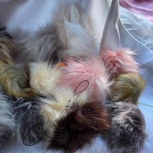 Può includere: Una collezione di pompon colorati e soffici con cordini neri attaccati. I pompon sono in varie tonalità di rosa, bianco, grigio e marrone.
