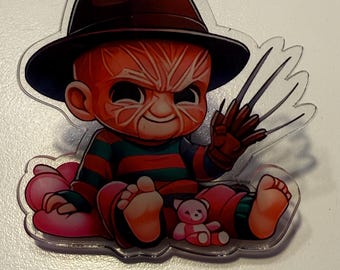 Puede incluir: Un charm de acrílico transparente que representa a Freddy Krueger bebé. El personaje lleva un sombrero marrón, una camisa a rayas rojas y verdes y pantalones rojos. Tiene un guante con garras y está sentado en una nube rosa con un osito de peluche.